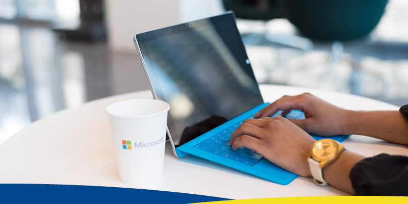 Notebook em cima de mesa, com copo ilustrado com o logotipo da Microsoft ao lado