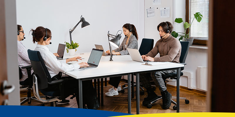 4 pessoas sentadas em uma mesa, em frente a notebooks e usando headsets
