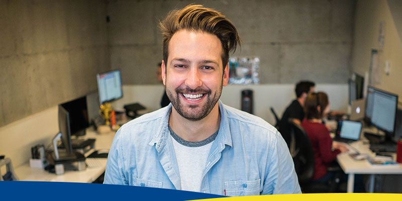 Homem olhando para frente, usando camisa azul, sorrindo, em sala semelhante a um escritório