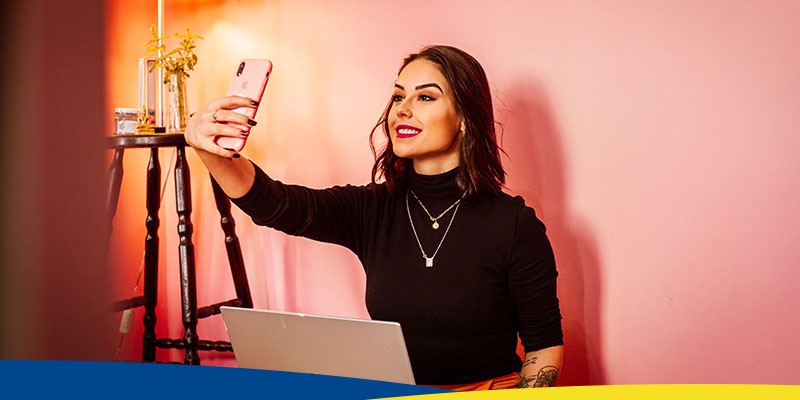 Mulher, de cabelos pretos, usando uma blusa preta, segura um celular com capa rosa em posição de selfie