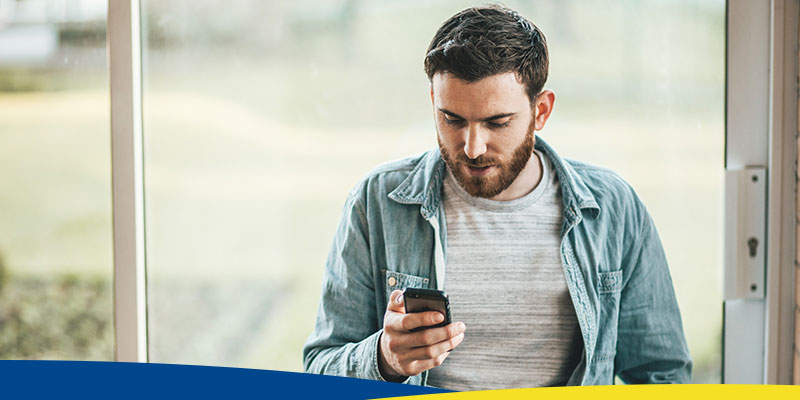 Homem, em frente a uma janela, olhando o celular
