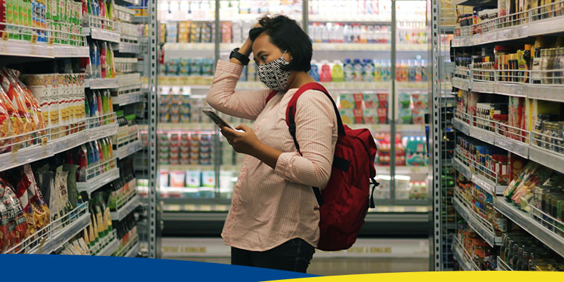 Mulher usando blusa listrada de mangas longas, branca e vermelha, e calça preta parada em frente a prateleiras de supermercado
