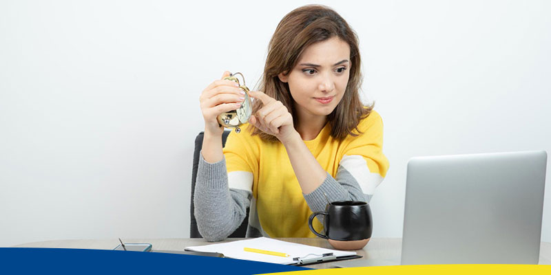 Foto de uma mulher, trabalhando em escritório, sentada em uma mesa segurando um relógio de ponteiros.